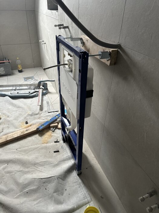 Inramad fixtur för vägghängd toalett monterad mot kakelvägg i badrum under renovering, med diverse verktyg och material runt omkring.