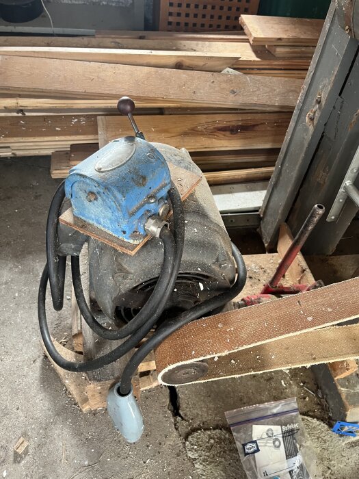 Äldre bandsåg med blå motor och rem, omgiven av träskivor, står på ett betonggolv.