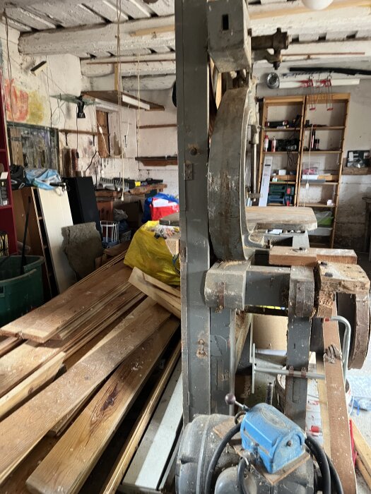 Äldre bandsåg i ett gammalt garage, omgiven av planks och verkstadsutrustning.