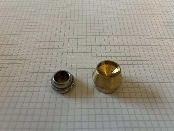 Två metallkomponenter ligger på ett rutat papper, en mindre och silverfärgad, en större och guldbrun.
