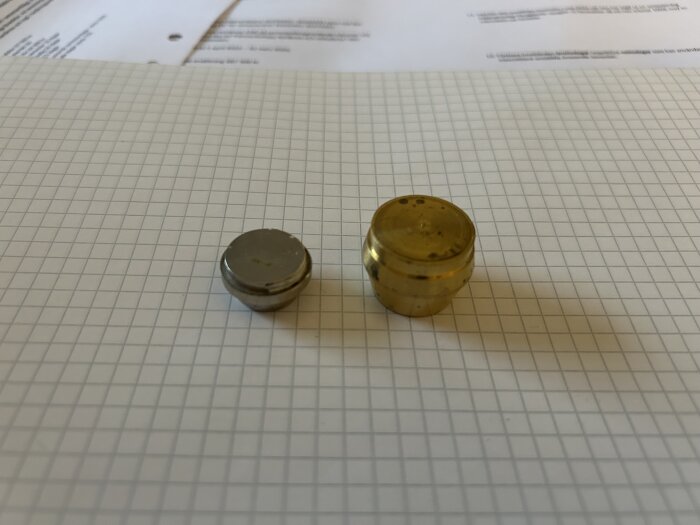 Två runda metallföremål, ett silverfärgat och ett mässingsfärgat, ligger på rutat papper med några dokument i bakgrunden.