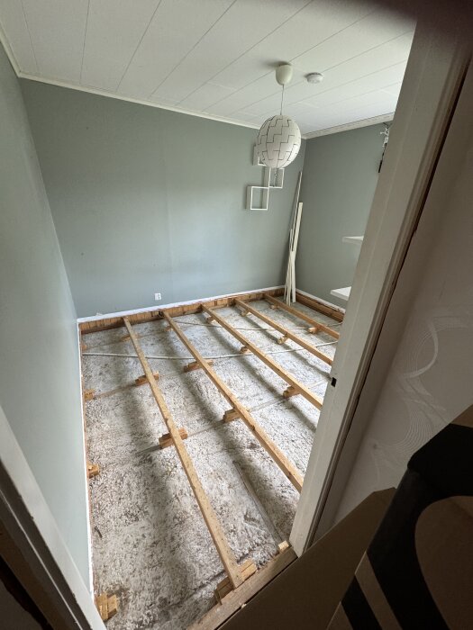 Rum med uppbrutna golv, synliga träreglar uppallade på träklossar över betongplatta, grå väggar och vitt tak med hängande lampa.