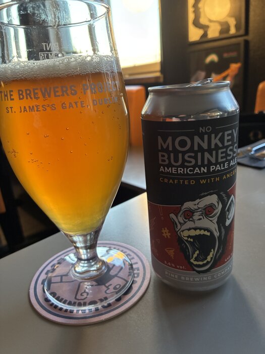 Glas med öl och en burk med etiketten "Monkey Business American Pale Ale" från Pine Brewing Company på ett bord.
