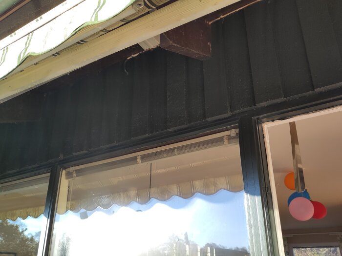Fasad och fönsterparti i ett hus, med en mörkmålad panel och en öppen markis ovanpå fönstret. Ballonger syns inomhus.