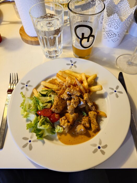 Tallrik med fläskfilégryta i sås, pommes frites, och sallad med grönsaker, serverad med dryck i glas.