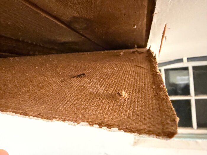 Brun och vit takplatta i nära bild, med fönster i bakgrunden. Eventuell diskussion om materialets ålder och asbestrisk.