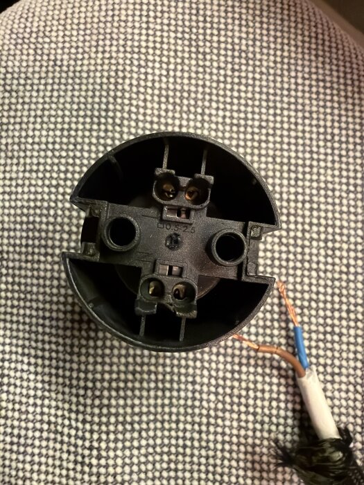 Elektrisk anslutning med två runda hål och synliga koppartrådar från en kabel, liggande på ett mönstrat underlag.