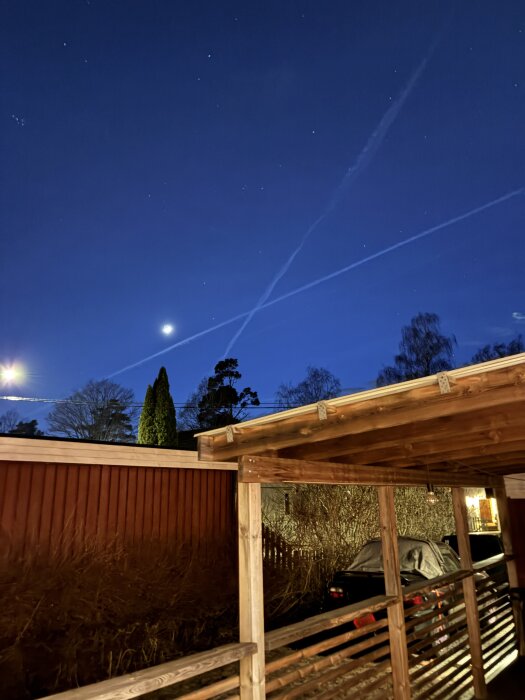 Nattbild av en träcarport och en delvis täckt bil, med en stjärnklar himmel och månen synlig ovanför med korsande flygplansspår i bakgrunden.