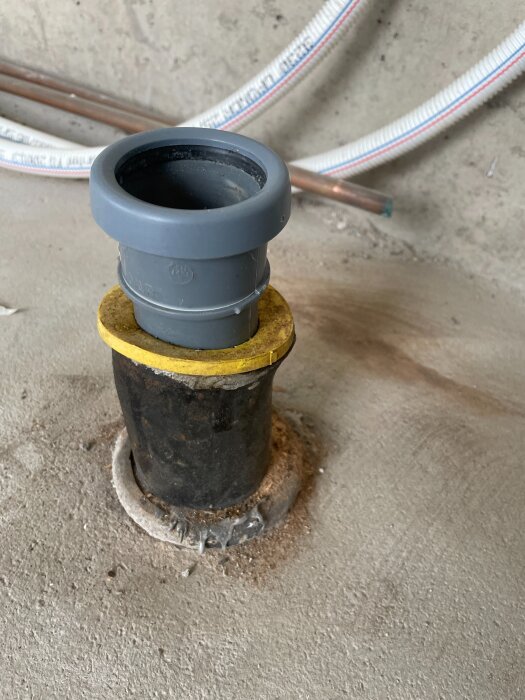 Grå plastförslutning på svart rör i betonggolv, med vita och kopparfärgade rör i bakgrunden.