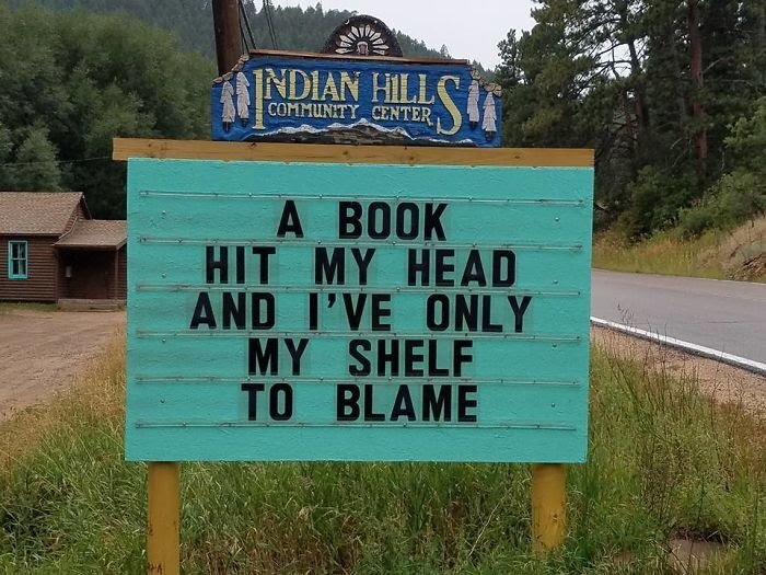 Skylt vid vägkant med text: "A book hit my head and I've only my shelf to blame" vid Indian Hills Community Center.