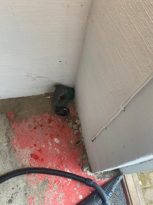 Håltagning i betongvägg för elmatning med borr och dammsugarsslang liggande på golvet, omgiven av grus och röd markering.