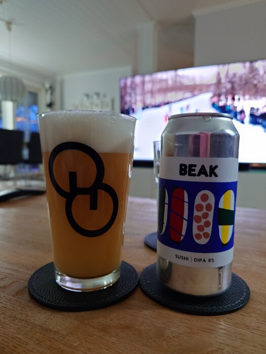 Glas med öl bredvid en burk med etiketten "BEAK" på ett bord, tv syns i bakgrunden.