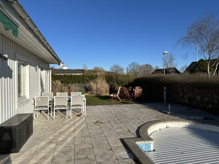 Terrass med matgrupp nära huset, pool i nederkant och häck i bakgrunden under klarblå himmel.