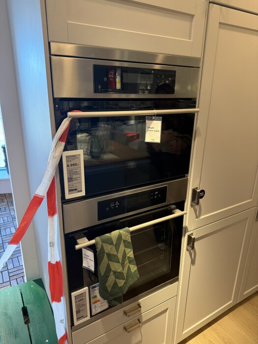 Inbyggd ugn i kök med rostfri skena och list, grön duk hänger på dörrhandtaget, etikett med prisinformation och text "Utgår inom kort".
