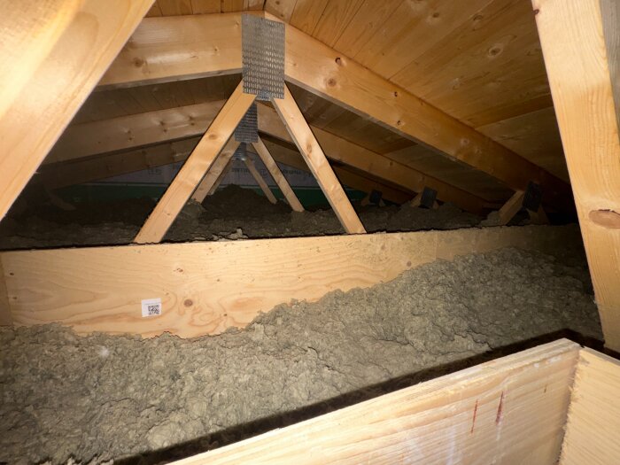 Vindsutrymme ovanför garage med träbjälkar och lös isolering, där någon har lagt material direkt på isoleringen.