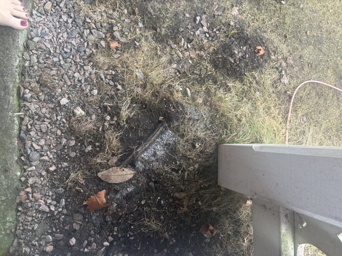 Rör i marken synligt ovan jord med frost på, omgiven av grus och gräs, intill en byggnad och en fot i nedre vänstra hörnet.