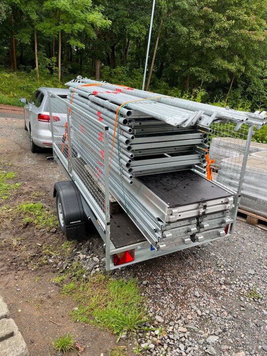 Stålställning på släpvagn vid bil, omgiven av grönt och grusplan. Förberett för husmålning och byggprojekt, i nyskick.
