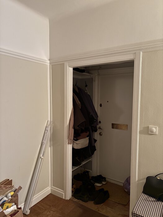 Ingång med dörr som öppnas mot en hall, fylld med skor och kläder. Vägg som kan komma att rivas markerad för renovering.