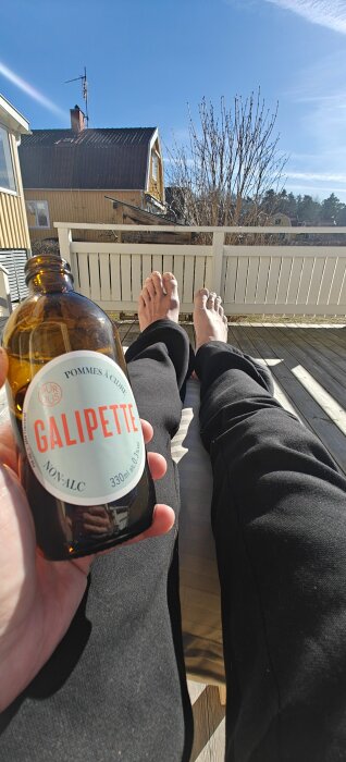 En person ligger på en solig altan, håller en alkoholfri ciderflaska med etiketten "Galipette", och vilar fötterna på en stol.