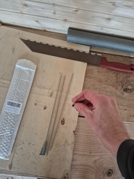 Hand som håller en fibertråd bredvid metallrör på ett bord, med en kniv och förpackning i bakgrunden.