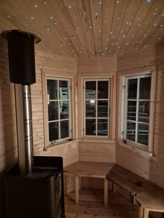 Bastuinredning med stjärnhimmel i taket, tre fönster och en kamin. Täta små ljuspunkter ger illusion av stjärnor i innertaket av trä.