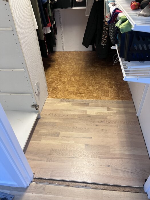 Nylagt parkettgolv i en klädkammare, delvis inrett med hyllor och kläder, med en tydlig tröskel mellan rummen.
