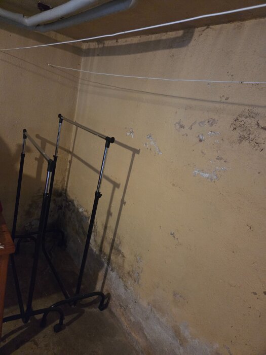 Sliten källarvägg med avflagnad färg, beiga tapeter, och två klädställningar utan kläder.