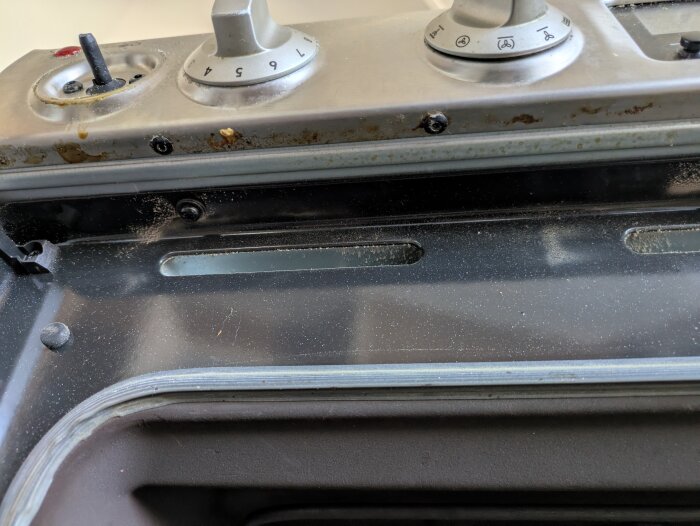 Stovetop knobs and screws on a stainless steel panel of an oven, with visible dirt and grime.