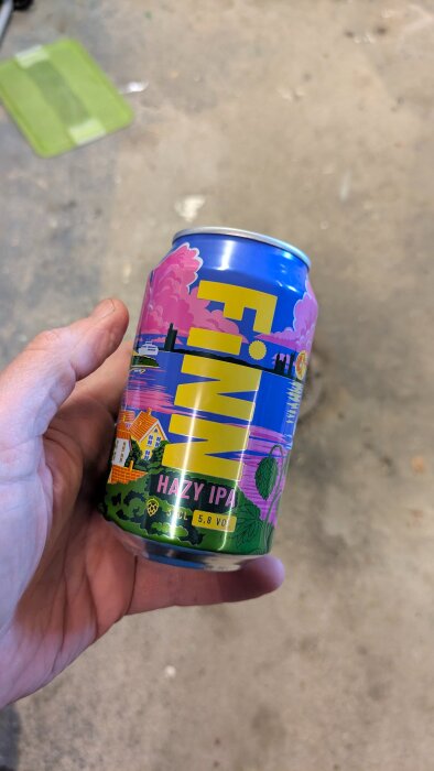 En person håller en burk med F.I.N.N. Hazy IPA, 5,8% volym. Bakgrund med färgglada illustrationer och landskapsmotiv.