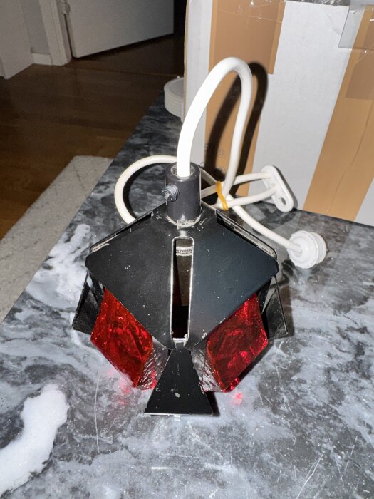Svart och röd lampa med exponerad kabel och skruvad plastdel på marmorbord, pappkartong i bakgrunden.