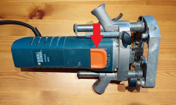 En gammal Black & Decker fräs, liggandes på ett träbord, med en röd pil som markerar strömbrytaren på sidan.