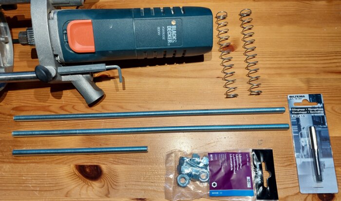 Elektrisk fräs från Black & Decker, tre gängstänger, muttrar, spiralfjädrar och en gängtapp från Biltema på ett träbord.