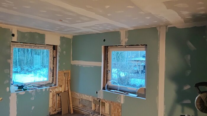 Rum under renovering med spacklade gipsskivor på väggar och tak, två stora fönster och byggmaterial framför fönstret.