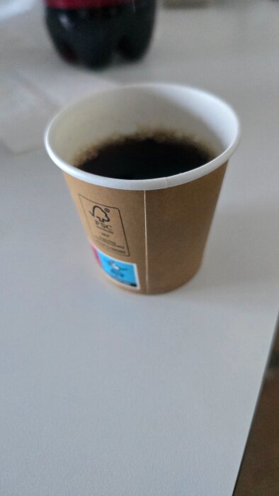 Pappersmugg med svart kaffe på ett vitt bord, en mörk dryckesflaska syns suddigt i bakgrunden.