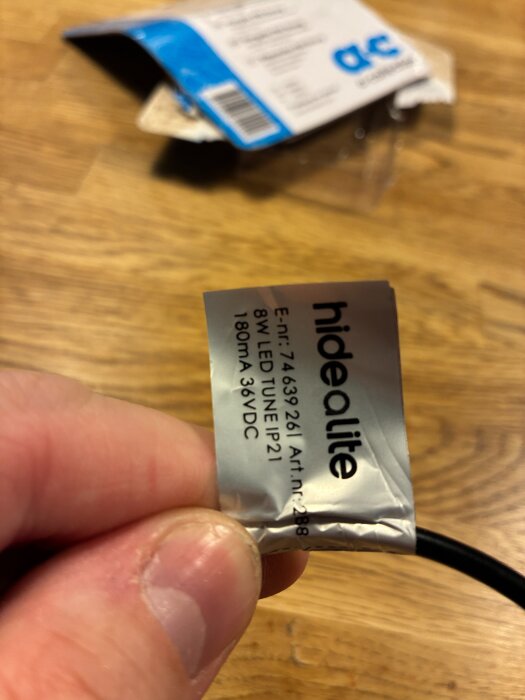 En etikett på en Hide a Lite driver, 8W LED TUNE IP21, 180mA 36VDC, hålls mellan fingrarna mot en suddig bakgrund med en förpackning.