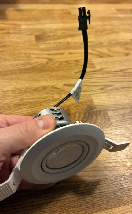 Hand håller en rund spotlampa med vit ram och svart sladd med kontakt, placerad mot en träyta.