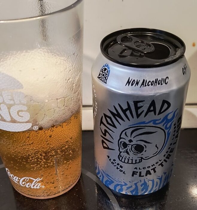 Burk med alkoholfri Pistonhead Flat Tire bredvid ett glas med gyllene dryck stående på ett bord.