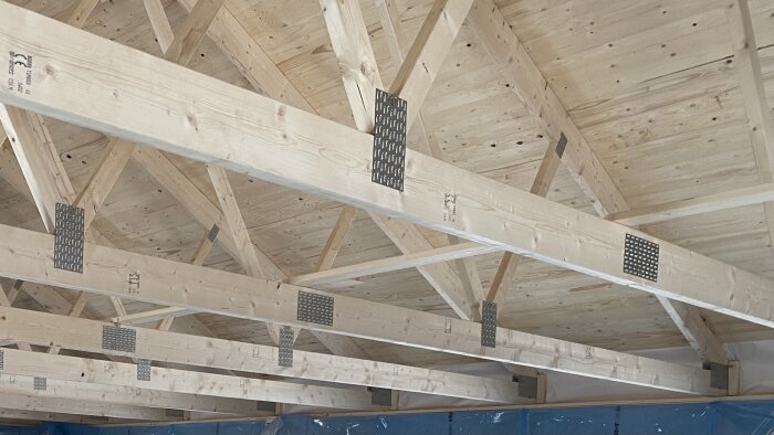 Takstolar i trä med metallplattor, sedda inifrån en byggnad. Detalj av takfoten diskuterad för fäste av terrassmarkis.