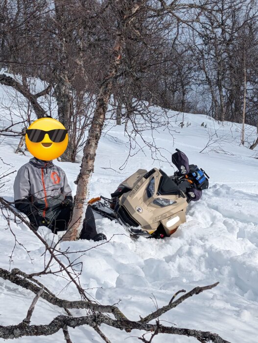 Snöskoter fast i snön vid fjällnära träd; person i vinterkläder sitter bredvid.
