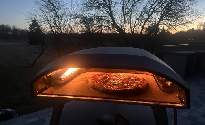 Pizza i en utomhusugn i solnedgången, svag eldslåga synlig i ugnen, omgiven av träd och skymning.