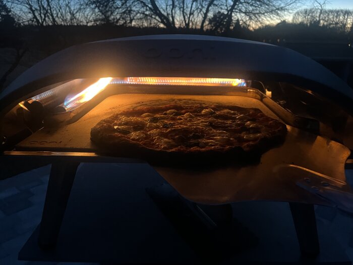 Pizza bakas i en ugn utomhus under skymning, med eld synlig i ugnen.