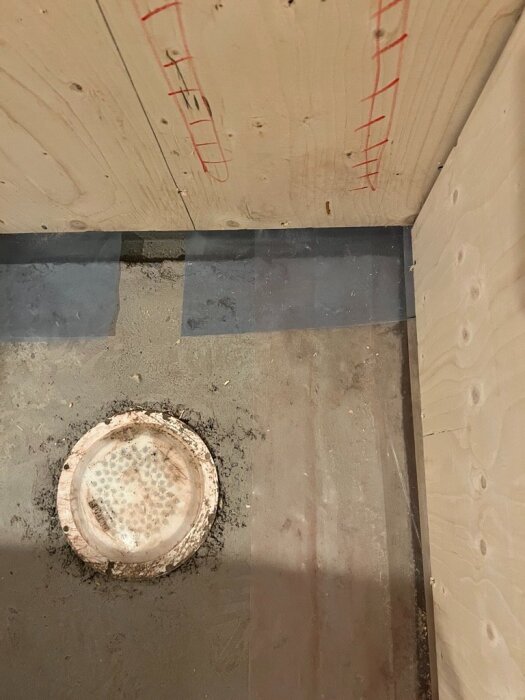 Badrumsgolv med betongplatta, plywoodskivor på väggarna och en urgröpning runt brunnen, förberett för gips och flytspackel.