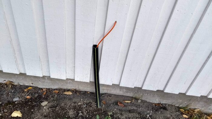 Svart plastskyddsrör med en orange kabel som sticker ut, lutande mot en vit trävägg, omgiven av jord och löv.