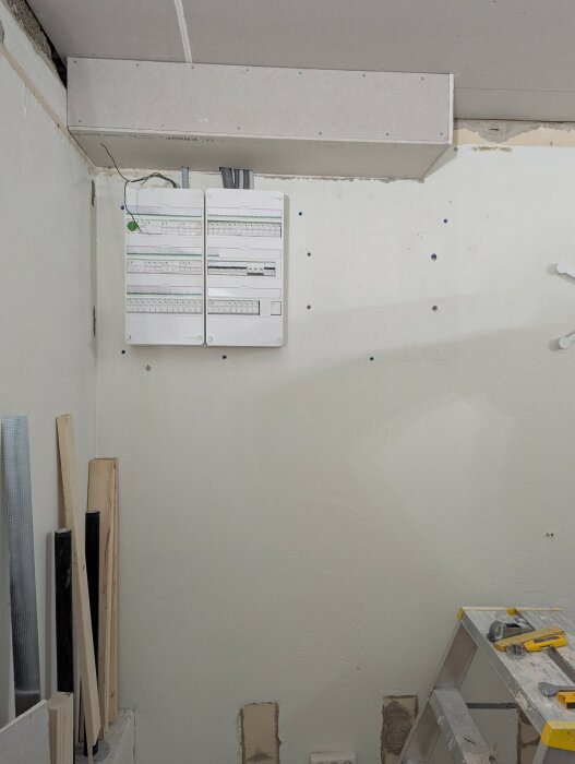 Elcentral monterad på en vit vägg med synliga hål för avlopp och elinstallationer. Verktyg och byggmaterial syns i hörnen.