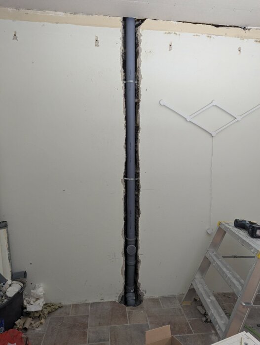 Inbyggt avloppsrör i en vit putsad vägg, med omgivande hålrum och byggmaterial i ett rum under renovering. Stege och verktyg syns.