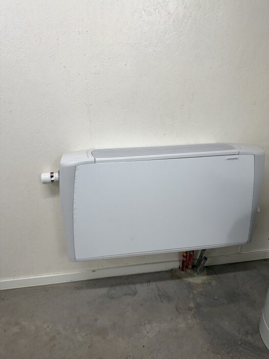 Bilden visar en vit väggmonterad radiator ovanför ett betonggolv med märken av fukt, bredvid syns några rör längs väggen.
