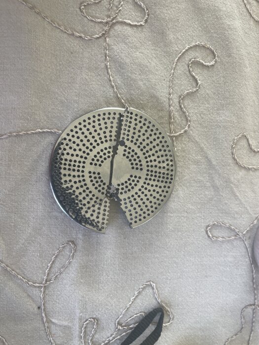 Rund metallplatta med hål och skåra, liggande på ett grått tyg med broderade mönster.