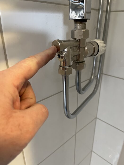En hand pekar på en hexskruv inuti en ventil kopplad till rör i ett badrum.