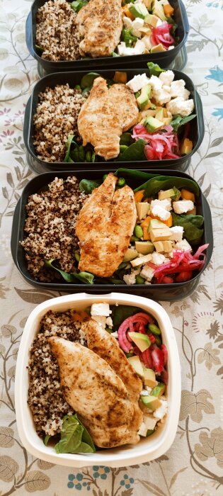 Matlådor med quinoa, kycklingfilé, spenat, sojabönor, fetaost, mango, avokado och picklad rödlök på ett blommigt bord.
