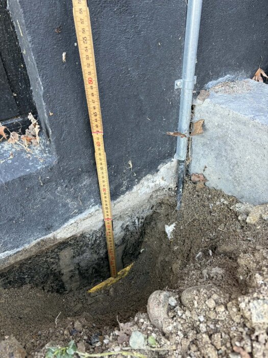 Grävd grop vid husvägg med mätsticka som visar 50 cm djup. Täcklist synlig nedanför marknivå. Mark och vägg utan tätning.